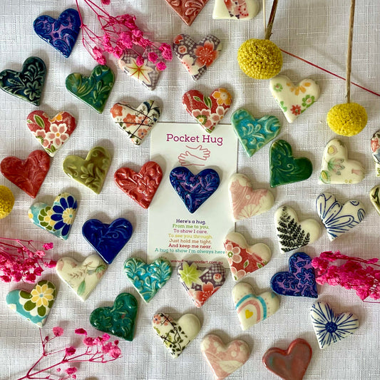 A collection of colorful, heart-shaped ceramic pocket hug ornaments with various floral and patterned designs, arranged on a white textured fabric background with a central white card that reads 'Pocket Hug'.