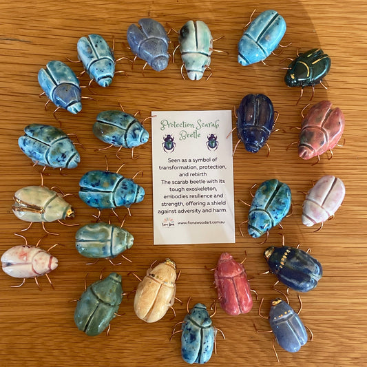 A circular arrangement of colorful beetle-shaped jewelry pieces on a wooden surface, with a small white card in the center reading 'Beetle Jewelry'. The jewelry includes blue, green, pink, beige, and dark blue pieces with metallic gold-toned details.