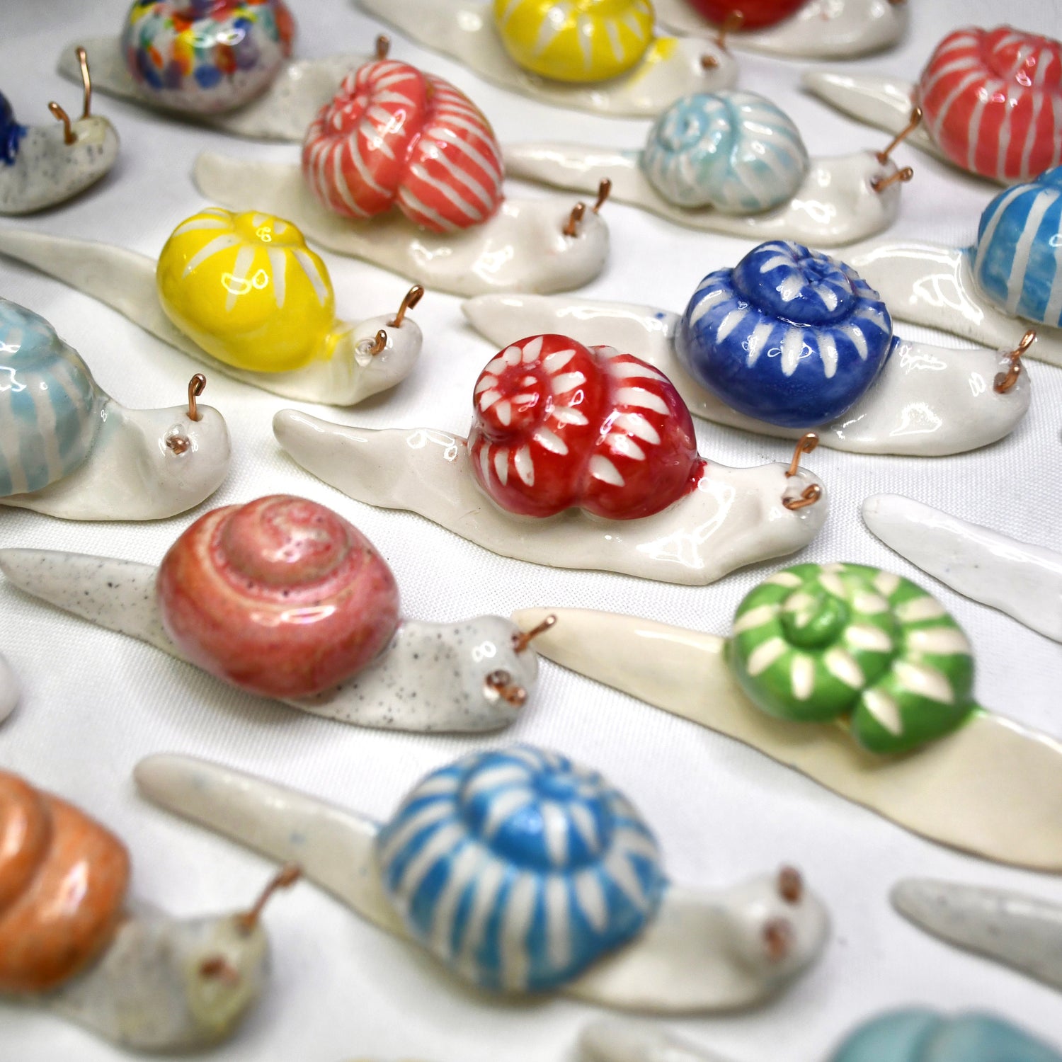 A close-up of multiple small ceramic snail ornaments arranged on a white surface. Each snail has a glossy shell in different colors including blue, red, yellow, green, pink, and orange, with cream-colored bodies and small metal hanging loops.