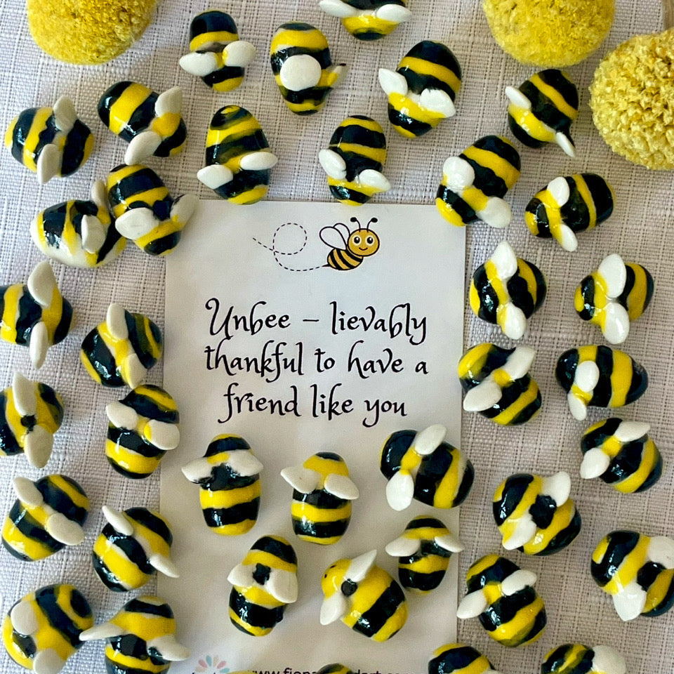 A collection of small bumblebee-shaped jewelry pieces arranged around a central printed card on a textured white fabric background. The card reads, 'Unbee - lievably thankful to have a friend like you' with a small bee illustration.