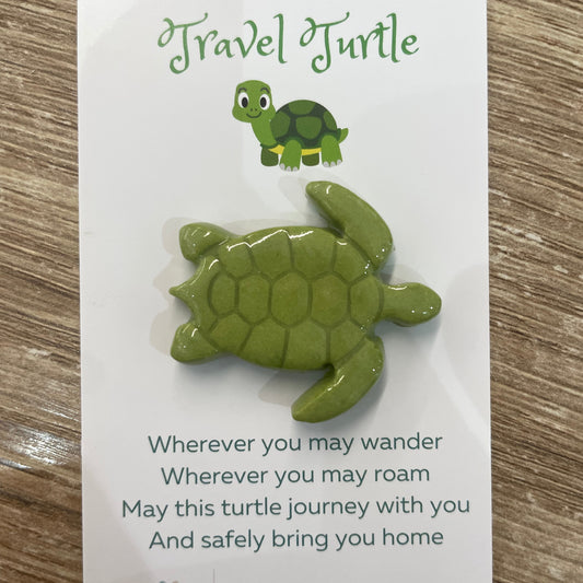 A small green turtle-shaped pin or charm displayed on a white backing card with printed text and a cartoon turtle logo, placed on a wood surface.