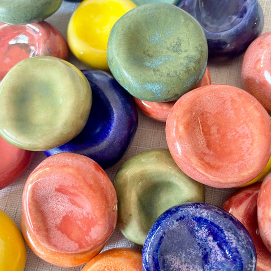 A close-up of multiple small, round, glossy ceramic dishes or bowls in assorted colors including blue, green, yellow, orange, pink, and purple, arranged on a light fabric surface.