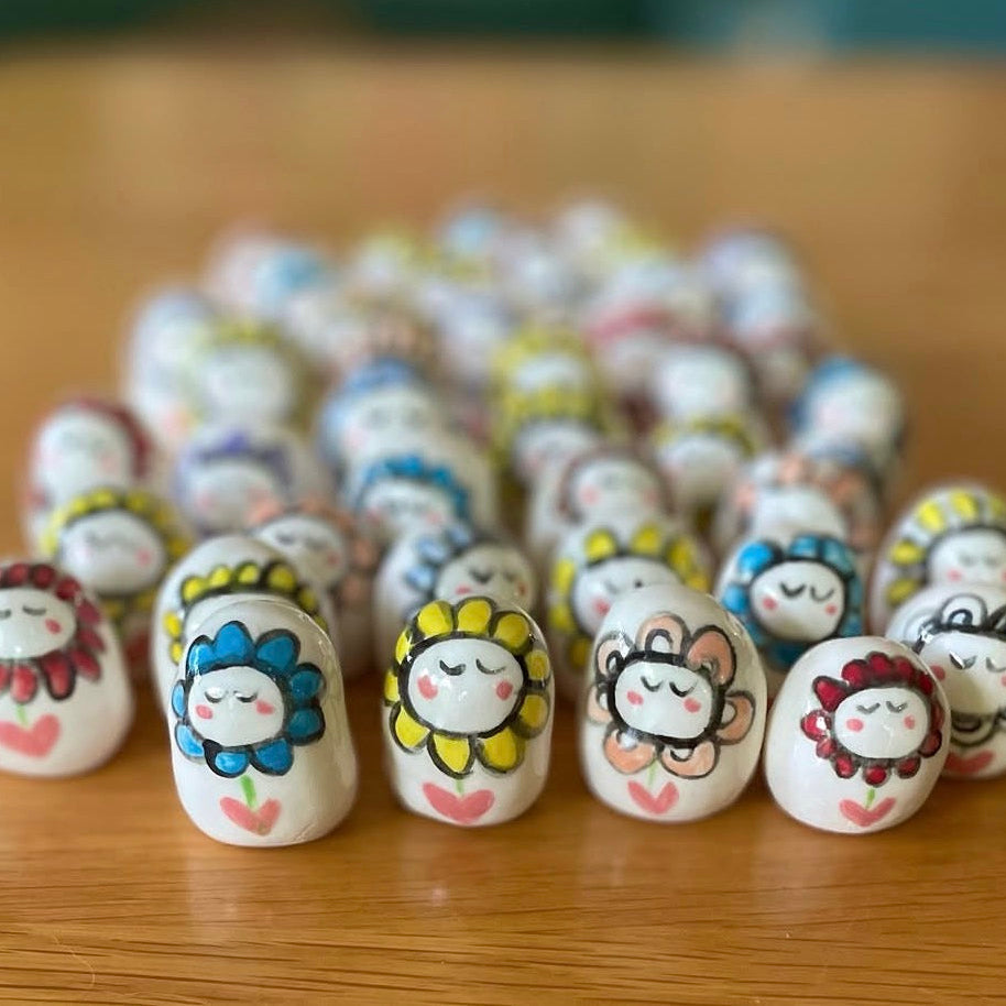 A group of small white ceramic beads or charms with hand-painted cartoon sunflower faces, arranged on a wooden surface.