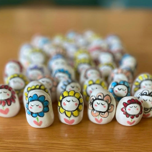 A group of small white ceramic beads or charms with hand-painted cartoon sunflower faces, arranged on a wooden surface.